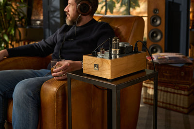 An incredibly attractive bearded male sits in a chair with a glass of whiskey listening to music on headphones and a Bottlehead Crack headphone amplifier.